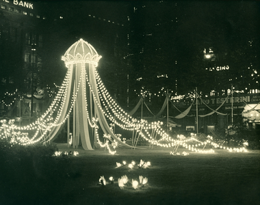 16949 - Nachtfoto van verlicht ornament met kroon en linten en sierlijke lampjes op de Dam in Amsterdam. Links op de ...