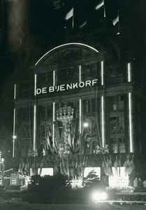 16950 - Nachtfoto van de Dam in Amsterdam met warenhuis De Bijenkorf. Op de voorgevel bevinden zich vlaggen en bloemen ...