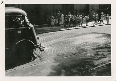 F006131 Kinderen kijken toe hoe de straat wordt schoongespoten m.b.v. de kolkenzuiger van de Plantsoen- en ...