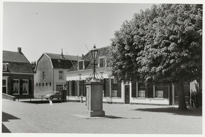  De oostzijde van de Brink met het gemeentehuis en de pomp en een grote boom vol blad. Links de brug en bebouwing aan ...