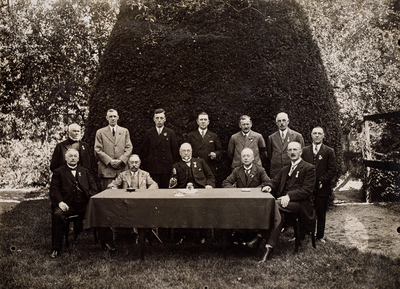  Groepsfoto van de gemeenteraad van Amerongen rond een tafel in een tuin, ter gelegenheid van het Oranjefeest