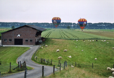  Onderaan de dijk de schuur met de afbeelding van een koe. Er landen 2 luchtballons met geel-rood gestreepte boven- en ...
