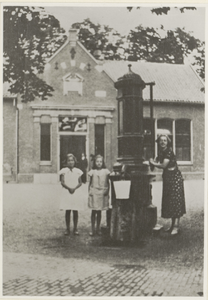  Twee meisjes en een mevrouw poserend bij de pomp op het Plein, op de achtergrond de voorgevel van de openbare lagere ...